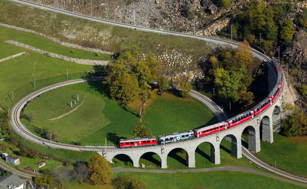 A red train crosses the stone-built spiral viaduct at Brusio, which loops in the open across a green meadow
