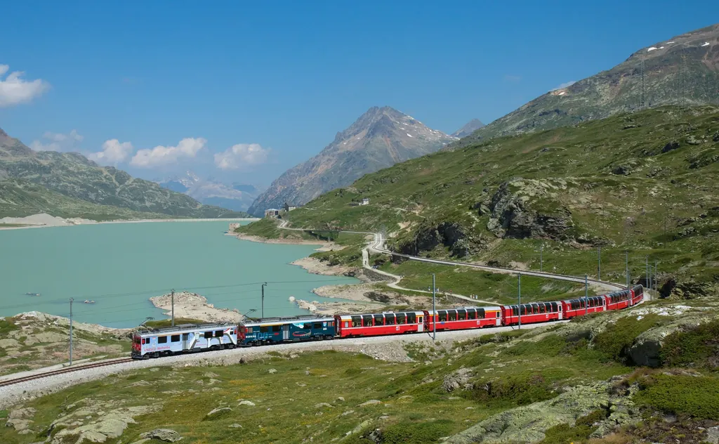 The Bernina Express with red panoramic cars runs along the shore of the turquoise Lago Bianco, with the peaks of the Bernina Pass behind
