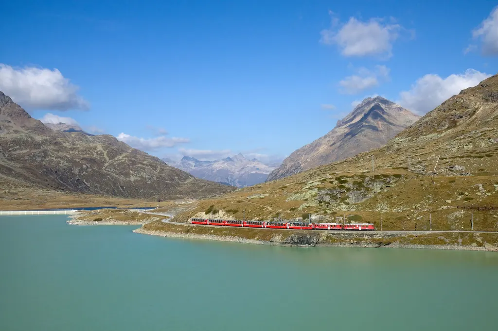 A red train runs along the shore of the milky turquoise Lago Bianco, surrounded by bare slopes under a blue sky