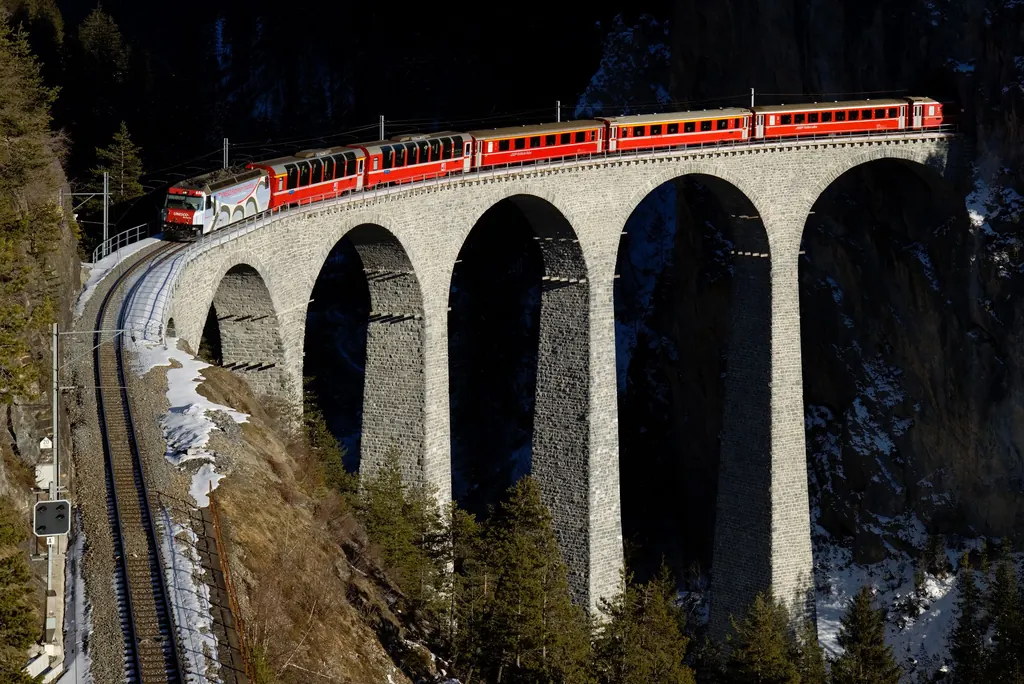 A red train with panoramic cars crosses the tall stone arches of the Landwasser Viaduct straight into a tunnel in the rock face