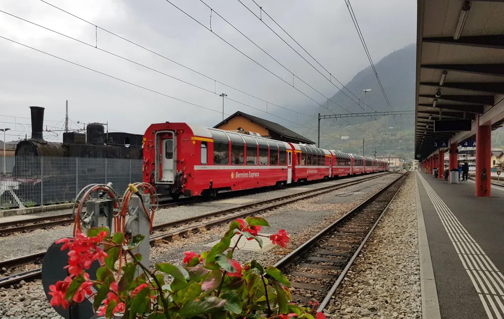 Rote Panoramawagen des Bernina Express mit bis ins Dach gezogenen Fenstern am Bahnsteig in Tirano, im Vordergrund rote Blumen
