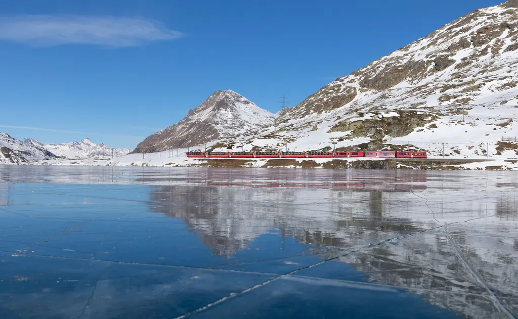 Der Bernina Express fährt am zugefrorenen, spiegelnden Lago Bianco entlang, dahinter verschneite Gipfel unter blauem Himmel