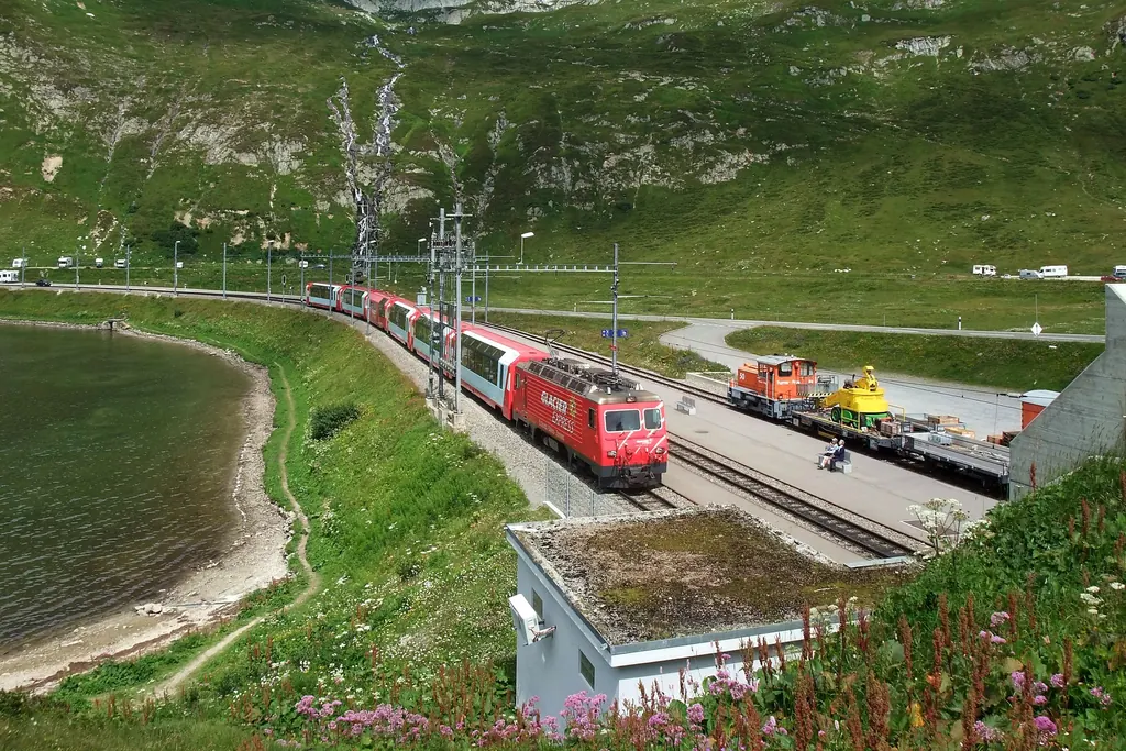 Der Glacier Express fährt im Sommer am Ufer des Oberalpsees entlang, im Vordergrund blühende Bergwiesen