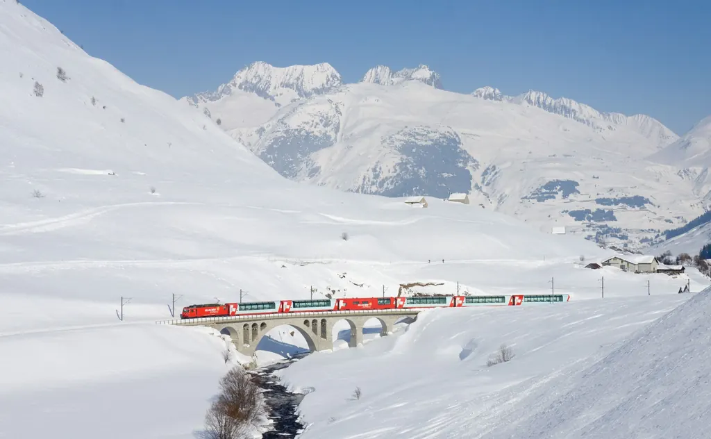 The Glacier Express crosses a stone bridge near Hospental, surrounded by deep-snow slopes and the peaks above the Urseren valley