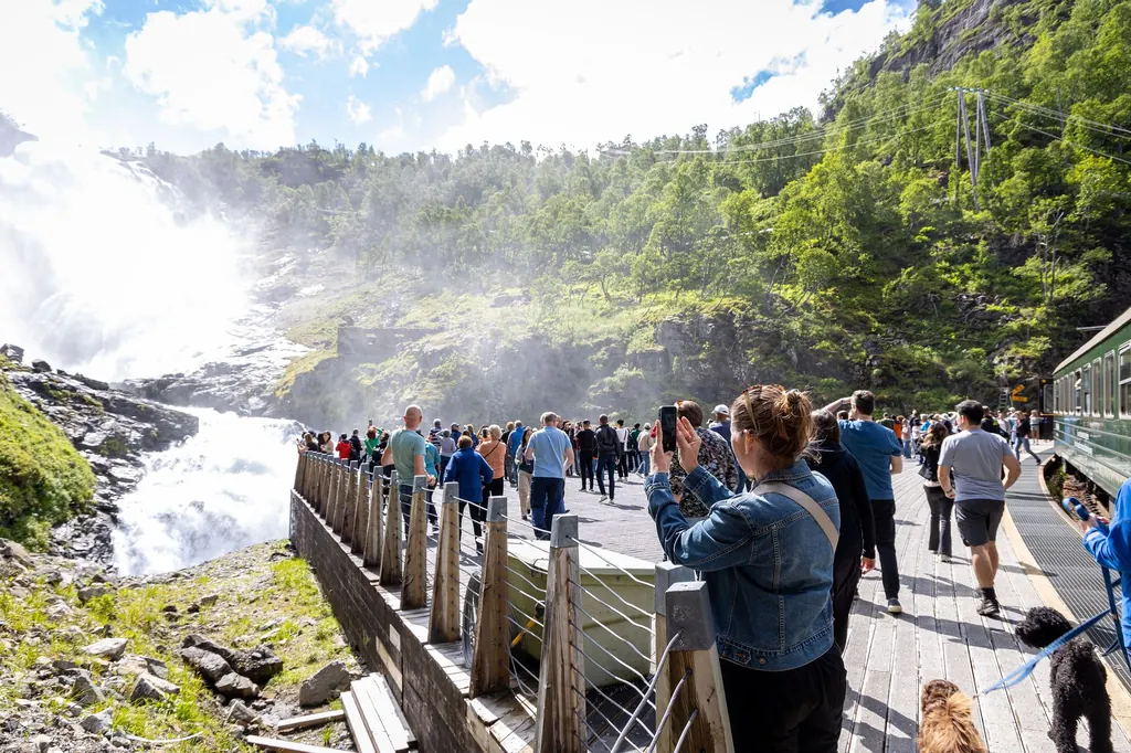 Fahrgäste stehen auf der hölzernen Plattform am Halt Kjosfossen und fotografieren den Wasserfall, rechts der grüne Zug