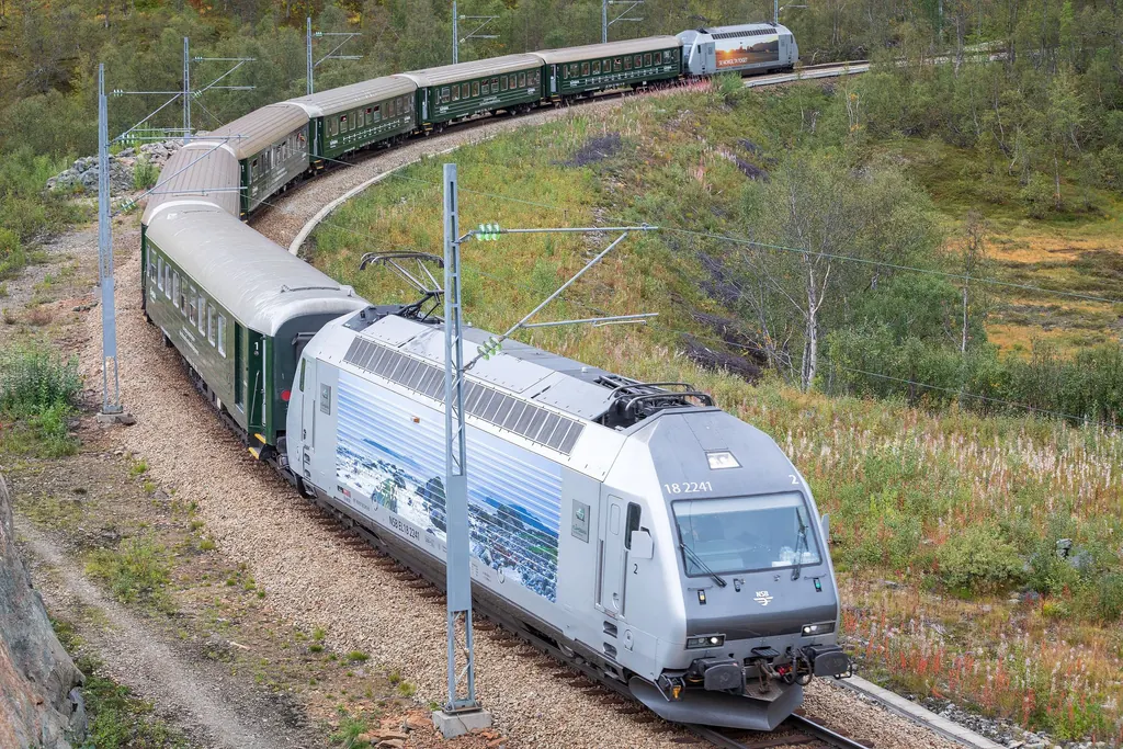Ein Zug der Flåmbahn mit silberner Lok und dunkelgrünen Wagen fährt durch eine enge Kurve am herbstlichen Berghang oberhalb des Flåmsdalen