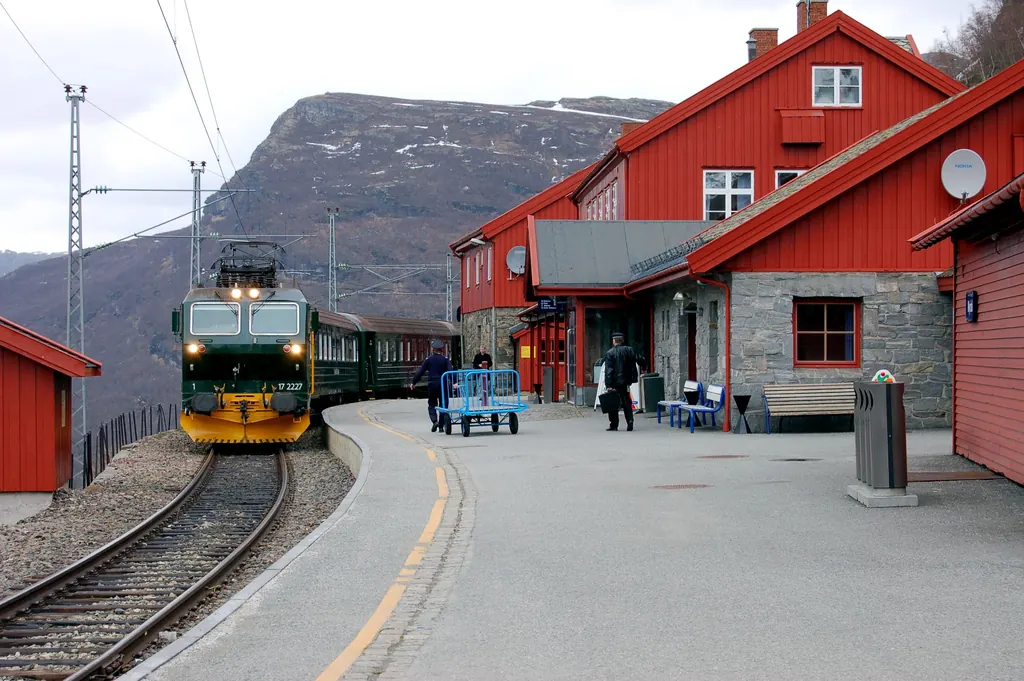 Ein grüner Zug der Flåmbahn steht am Bahnsteig von Myrdal neben den roten Holzgebäuden des Bahnhofs, dahinter kahle Berge mit Schneeresten