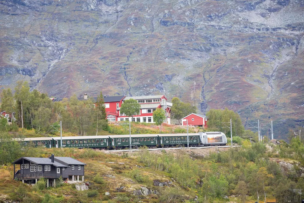 Ein Zug der Flåmbahn hält unterhalb des roten Berghotels Vatnahalsen, dahinter eine graue Felswand mit Wasserrinnen