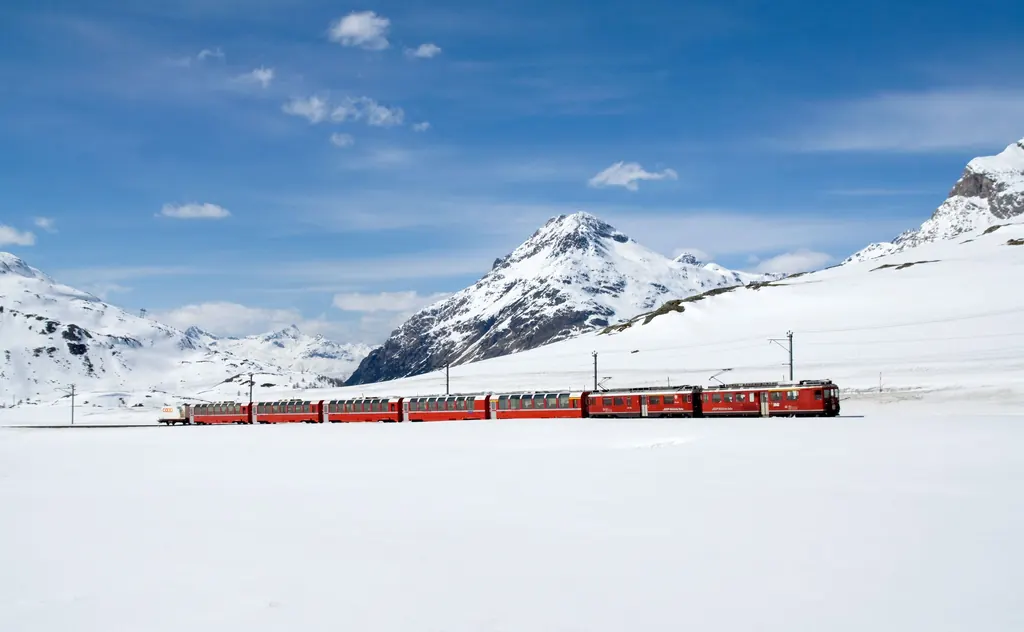 Ein roter Zug der Berninabahn fährt im Winter durch die tief verschneite Ebene am Lej Pitschen, dahinter weiße Gipfel unter blauem Himmel
