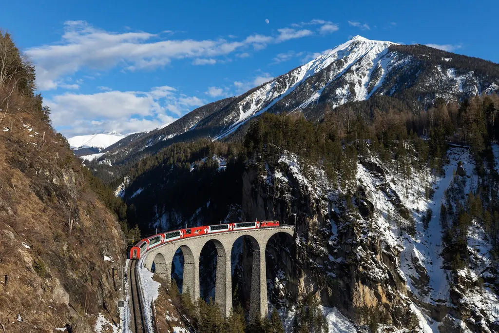 Ein Panoramazug fährt über den gemauerten Landwasserviadukt in den Tunnel in der Felswand, dahinter ein verschneiter Gipfel