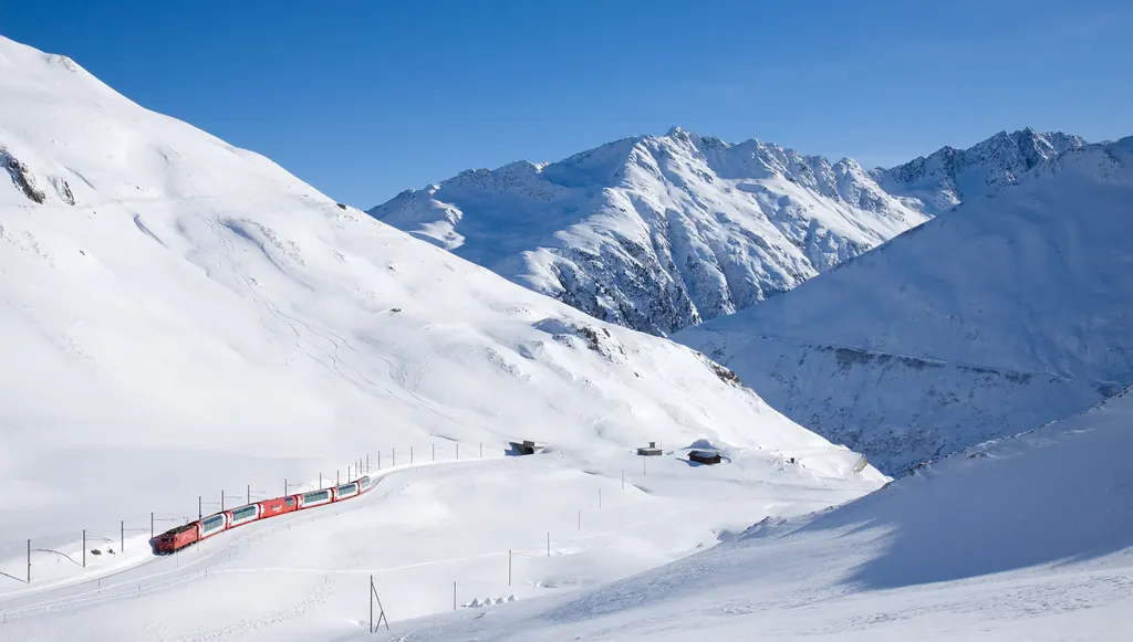 Der rote Glacier Express fährt klein durch eine weite, tief verschneite Berglandschaft kurz vor der Passhöhe des Oberalppasses