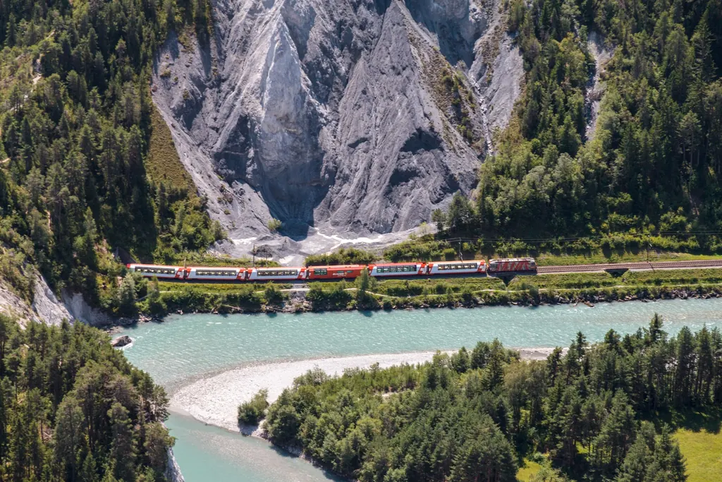 Der Glacier Express fährt am türkisfarbenen Vorderrhein entlang durch die Rheinschlucht, dahinter helle Erosionswände und Wald
