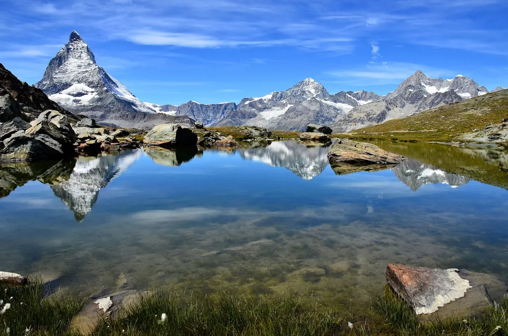 Das Matterhorn spiegelt sich im glatten Wasser des Riffelsees, im Vordergrund Steine und Gras, dahinter eine Kette verschneiter Gipfel