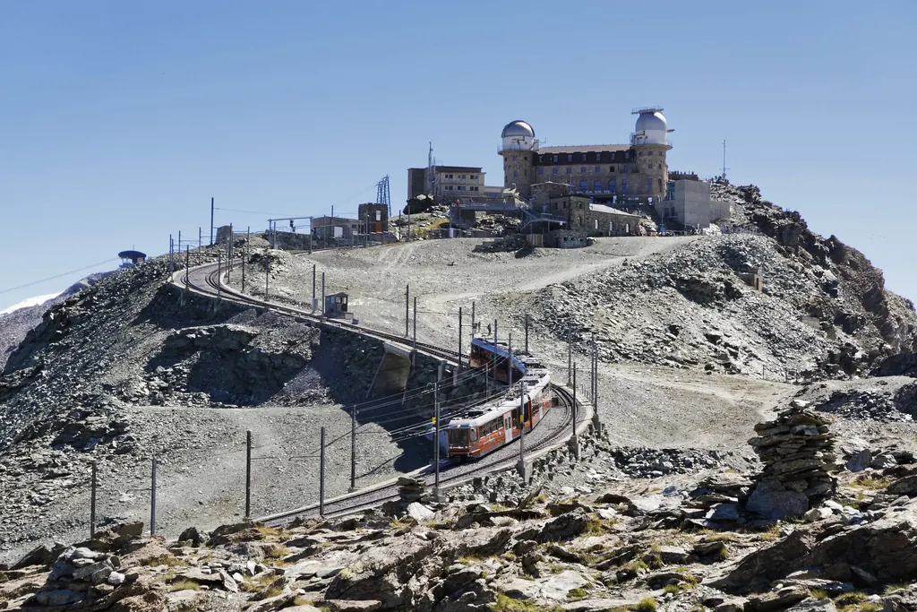 Ein Zug der Gornergrat Bahn fährt durch die letzte Kurve aus Geröll zur Bergstation, darüber das Kulmhotel mit seinen beiden Observatoriumskuppeln