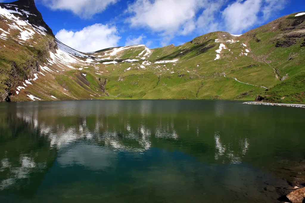 Der Bachalpsee liegt still zwischen grünen Hängen, im Wasser spiegeln sich Schneefelder und Wolken