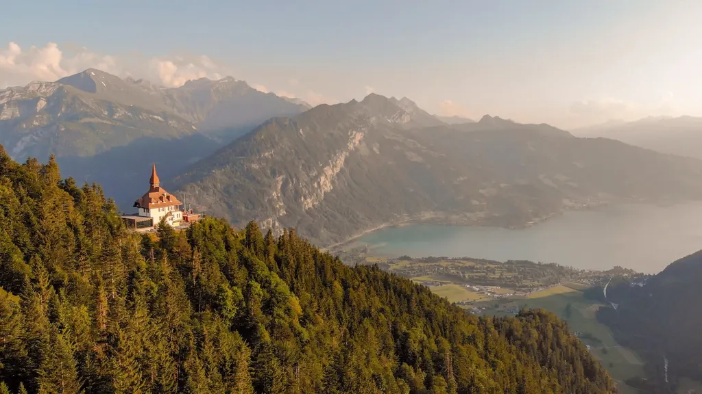 The Harder Kulm restaurant with its red turret roof on a wooded ridge in the evening light, Lake Thun and hazy mountain ranges behind it