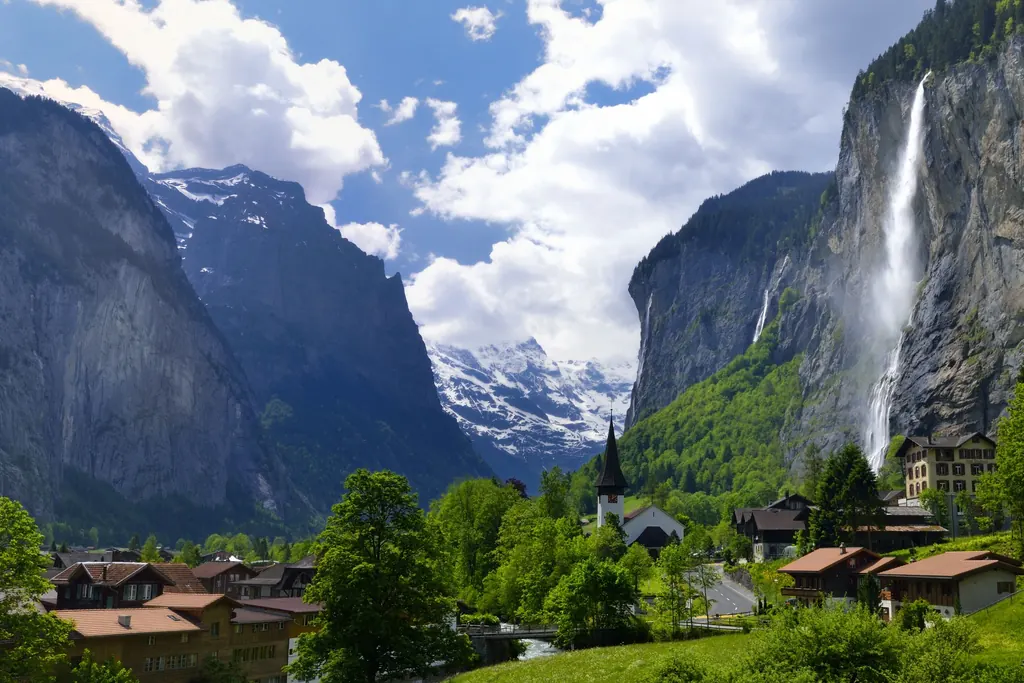 Das Dorf Lauterbrunnen mit Kirche und Holzhäusern im Talgrund, rechts stürzt der Staubbachfall über eine senkrechte Felswand, im Hintergrund verschneite Gipfel