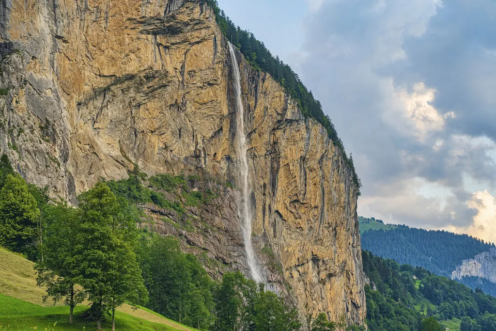 Der Staubbachfall fällt als dünner weißer Schleier über eine ockerfarbene, senkrechte Felswand, unten Bäume und Wiese