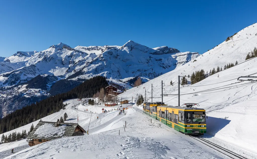 A green and yellow Wengernalp Railway train running through the snow below Kleine Scheidegg, snow-covered mountains behind it
