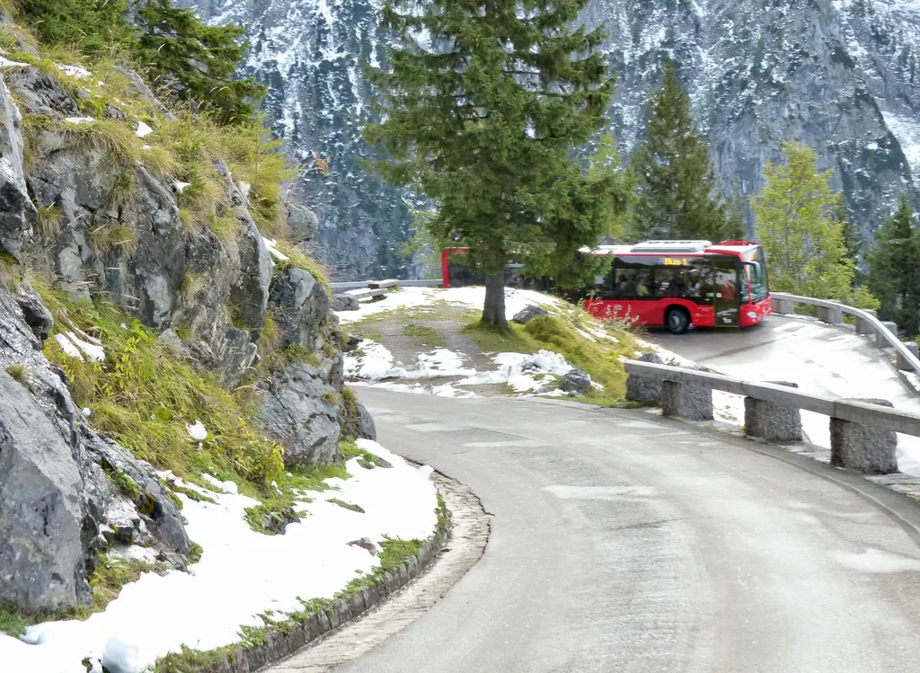 Ein roter Linienbus in einer Kurve der schmalen Kehlsteinstraße zwischen Fels, Lärchen und Schneeresten