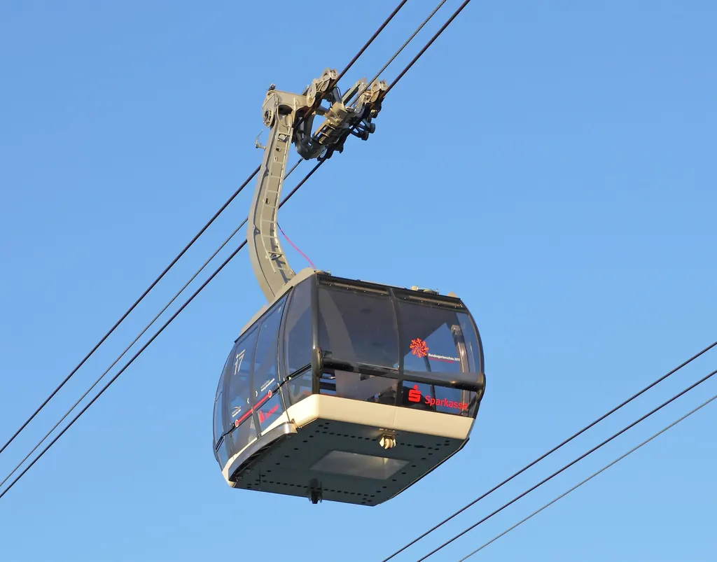 A cabin of the Koblenz cable car hangs from two track ropes against a blue sky, its seats arranged facing the windows