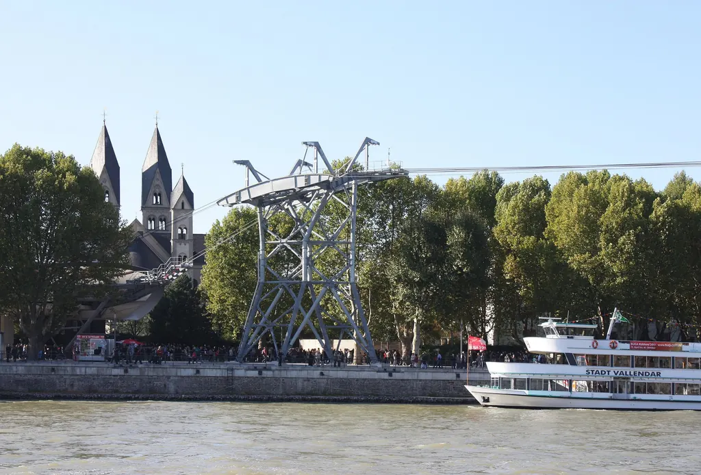 The valley station of the cable car on the bank of the Rhine with its steel structure, the two towers of St Castor's Basilica behind, an excursion boat in front