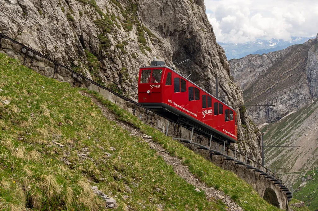 Ein roter Triebwagen der Pilatusbahn klettert die 48 Prozent steile Strecke hinauf, links grüne Wiese, rechts Fels, im Hintergrund die Berge