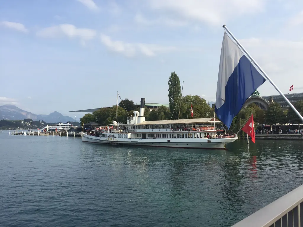 The paddle steamer Uri docks at the quay in front of Lucerne railway station, a Lucerne flag in the foreground, Lake Lucerne behind