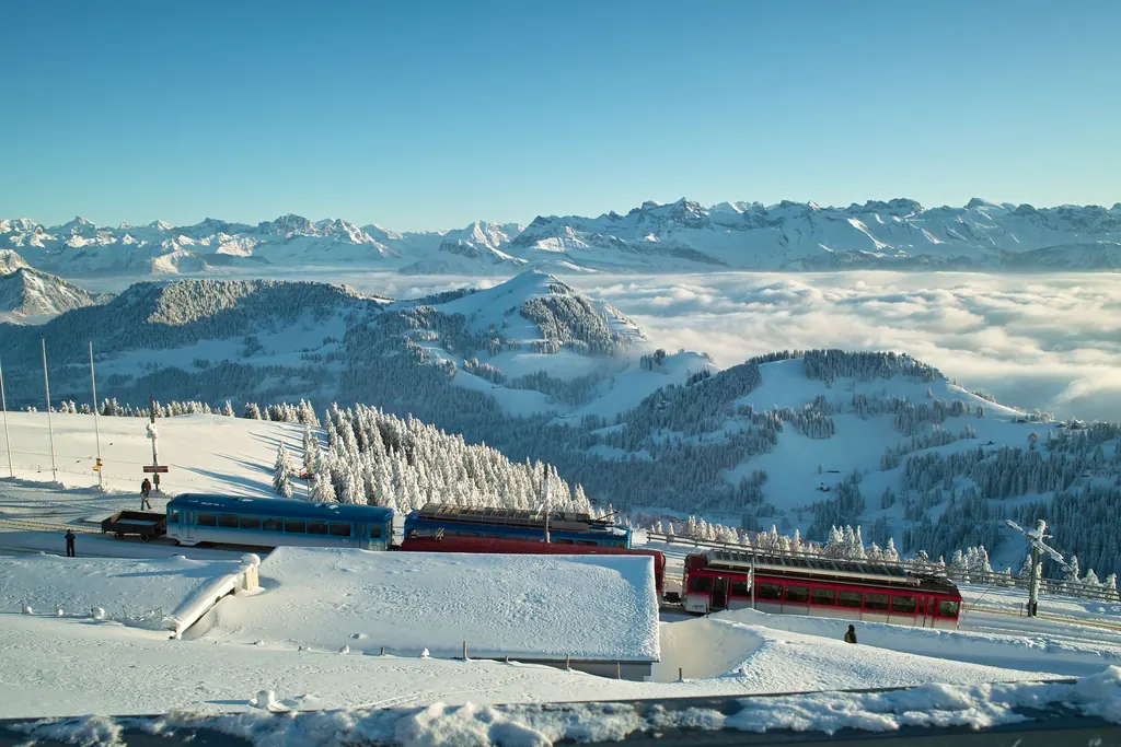 Rote und blaue Züge der Rigi-Bahnen im Schnee auf Rigi Kulm, dahinter ein Nebelmeer und die Alpen