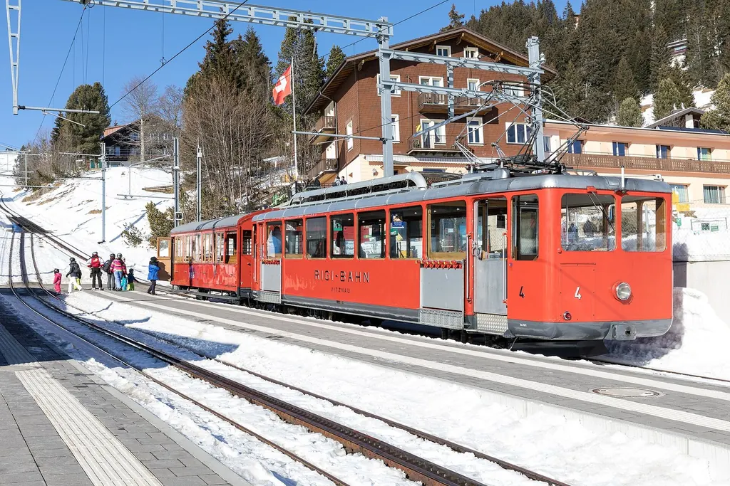 Ein roter Zug der Vitznau-Rigi-Bahn steht im Winter am Bahnhof Rigi Kaltbad, Schnee auf den Gleisen, dahinter ein Hotel