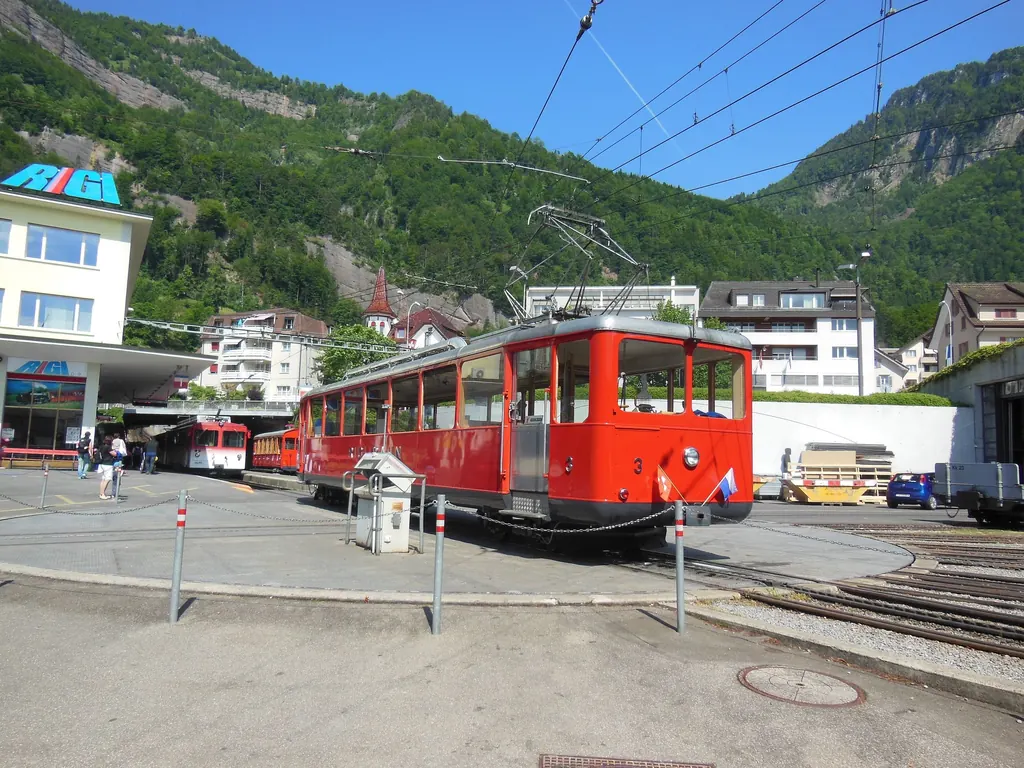 A red railcar of the Vitznau–Rigi railway stands at the Vitznau valley station, the wooded slopes of the Rigi behind