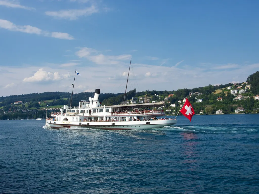 The paddle steamer Stadt Luzern with a Swiss flag at the stern crosses the blue Lake Lucerne, houses and woods on the shore